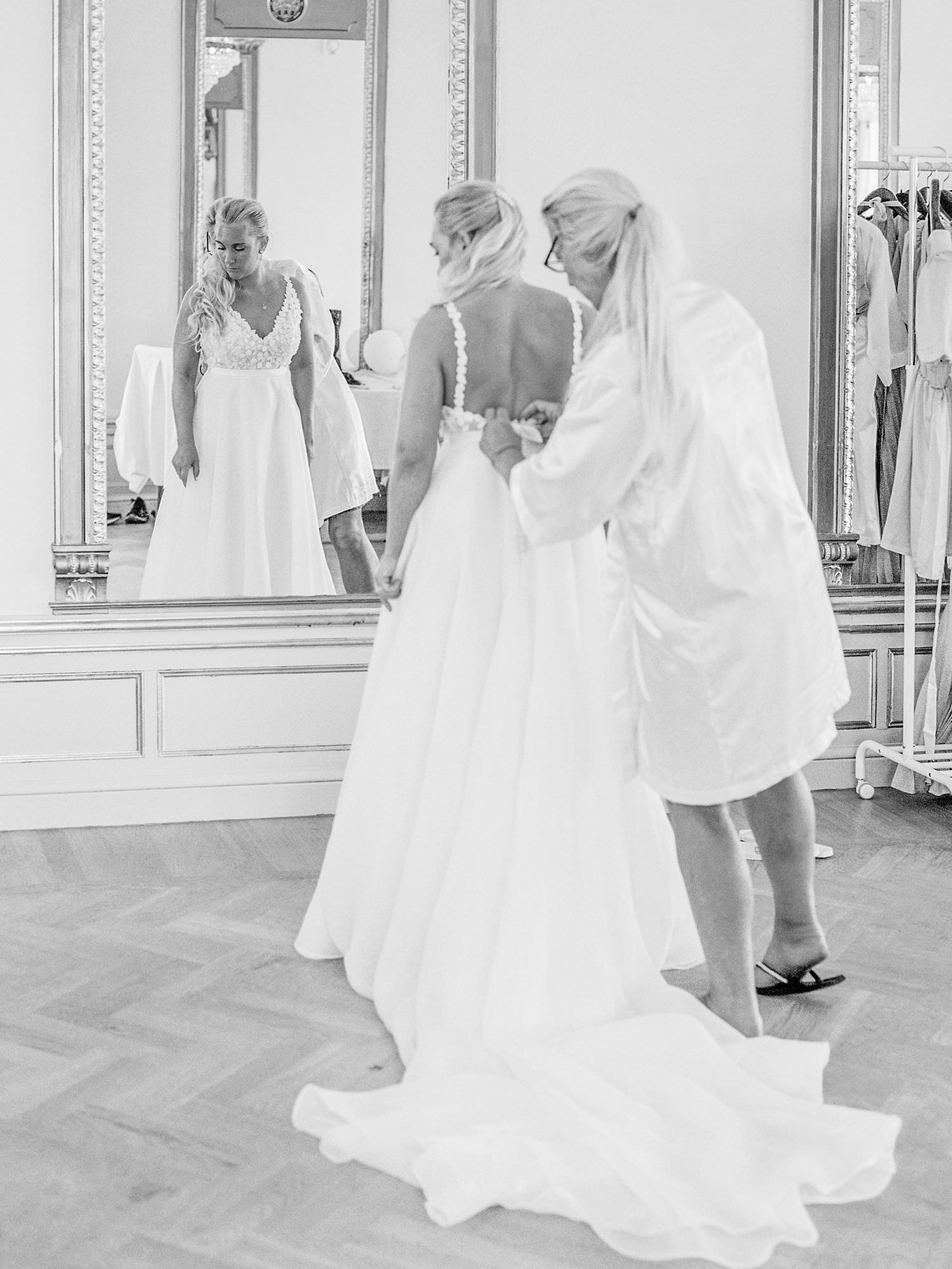 Black and white photograph of the bride getting ready. Her mother is helping her button the dress