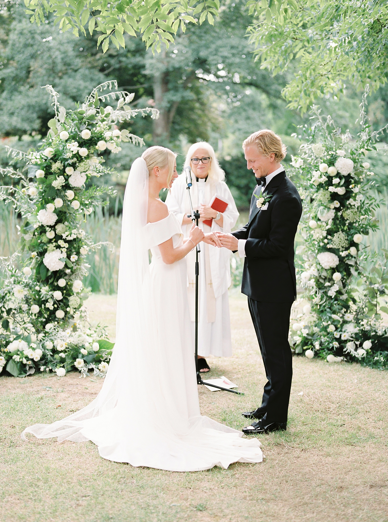 bride and groom exchange rings in front of a large floral arch filled with white flowers and greenery