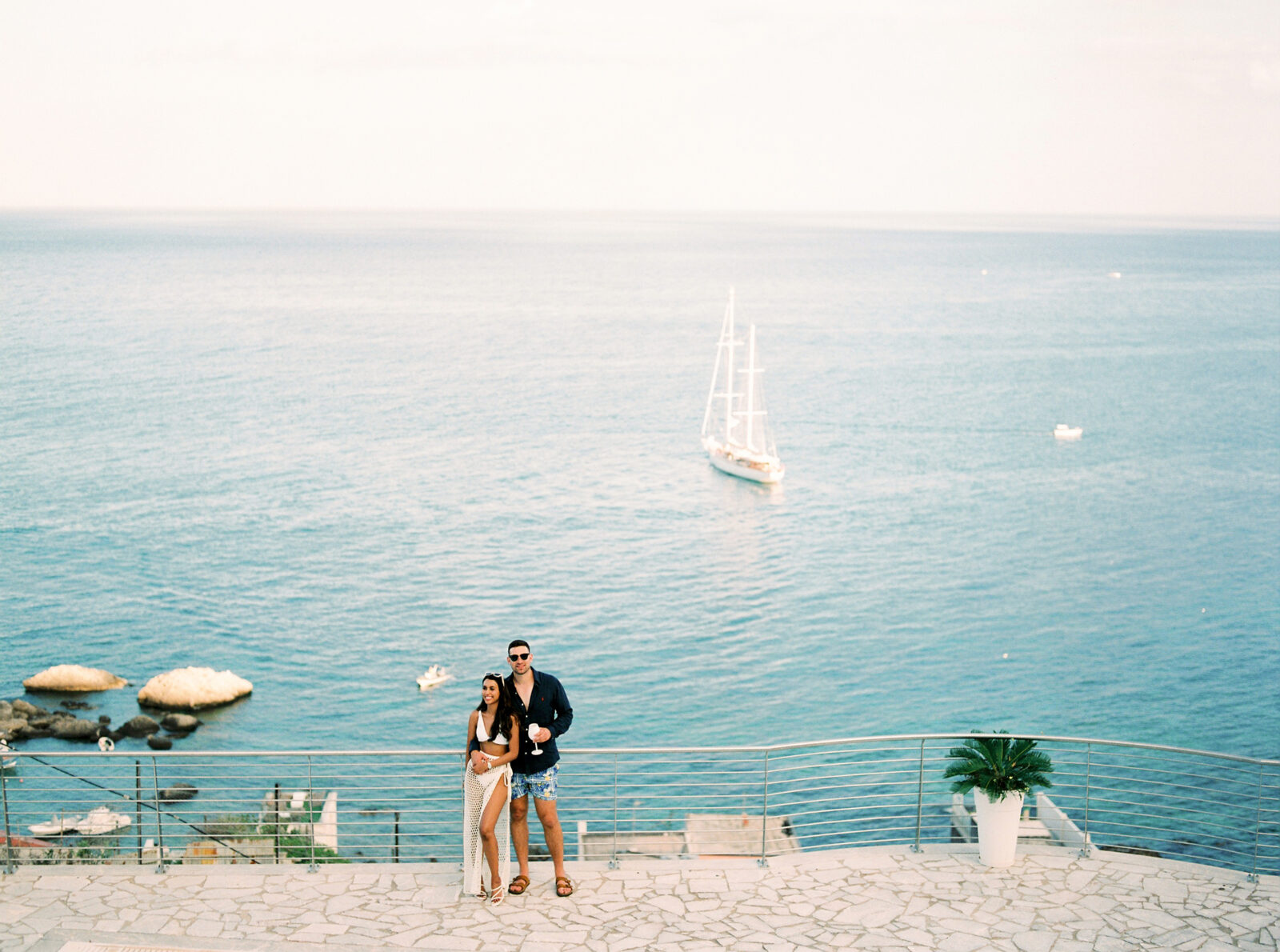 A couple standing on a seaside promenade with a sailboat in the background, overlooking the ocean at Baya Bella Taormina luxury villa.
