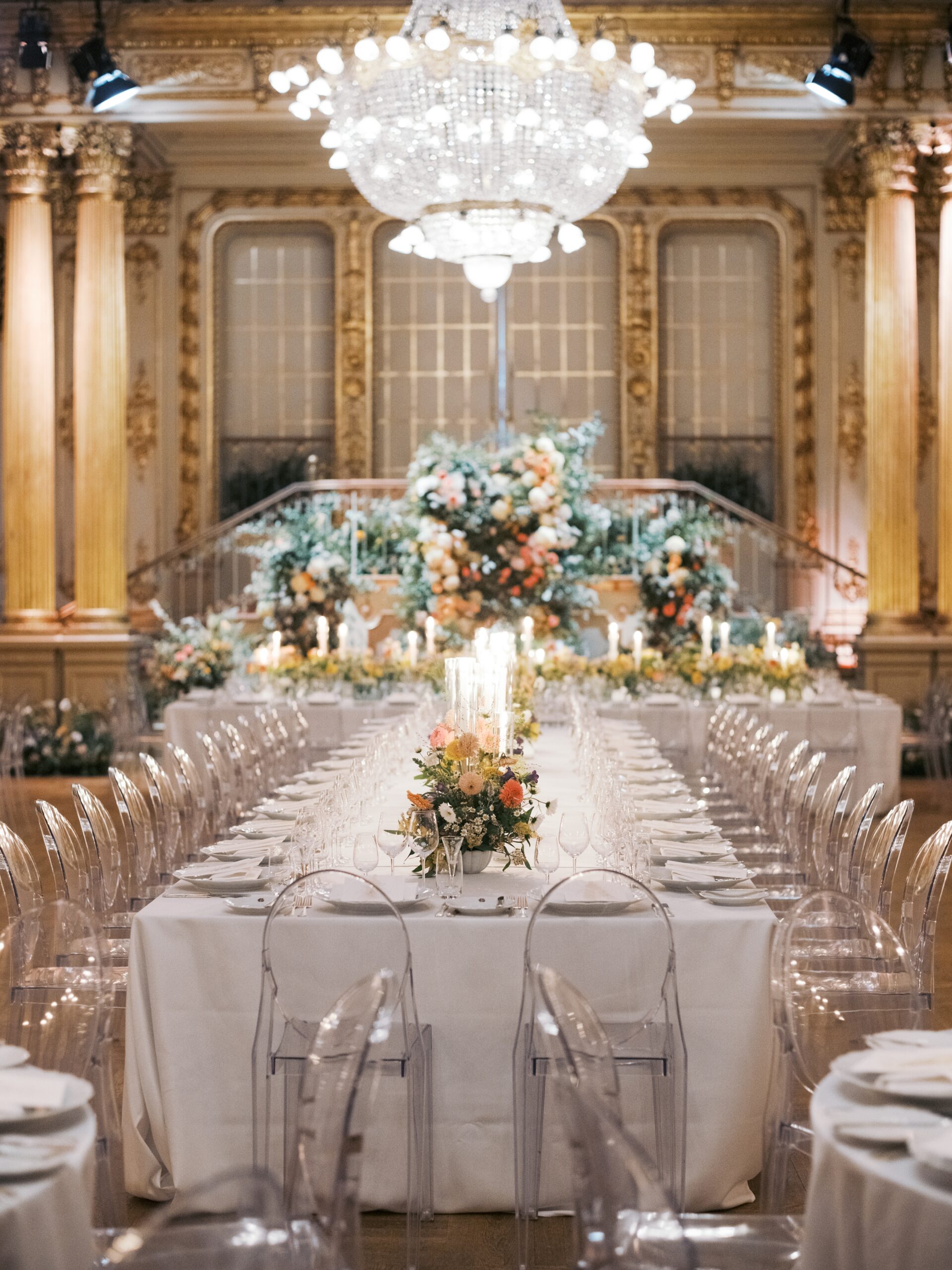 A candlelit wedding reception setup at Grand Hôtel’s Spegelsalen, featuring transparent chairs, long tables, and lush centerpieces beneath ornate chandeliers.