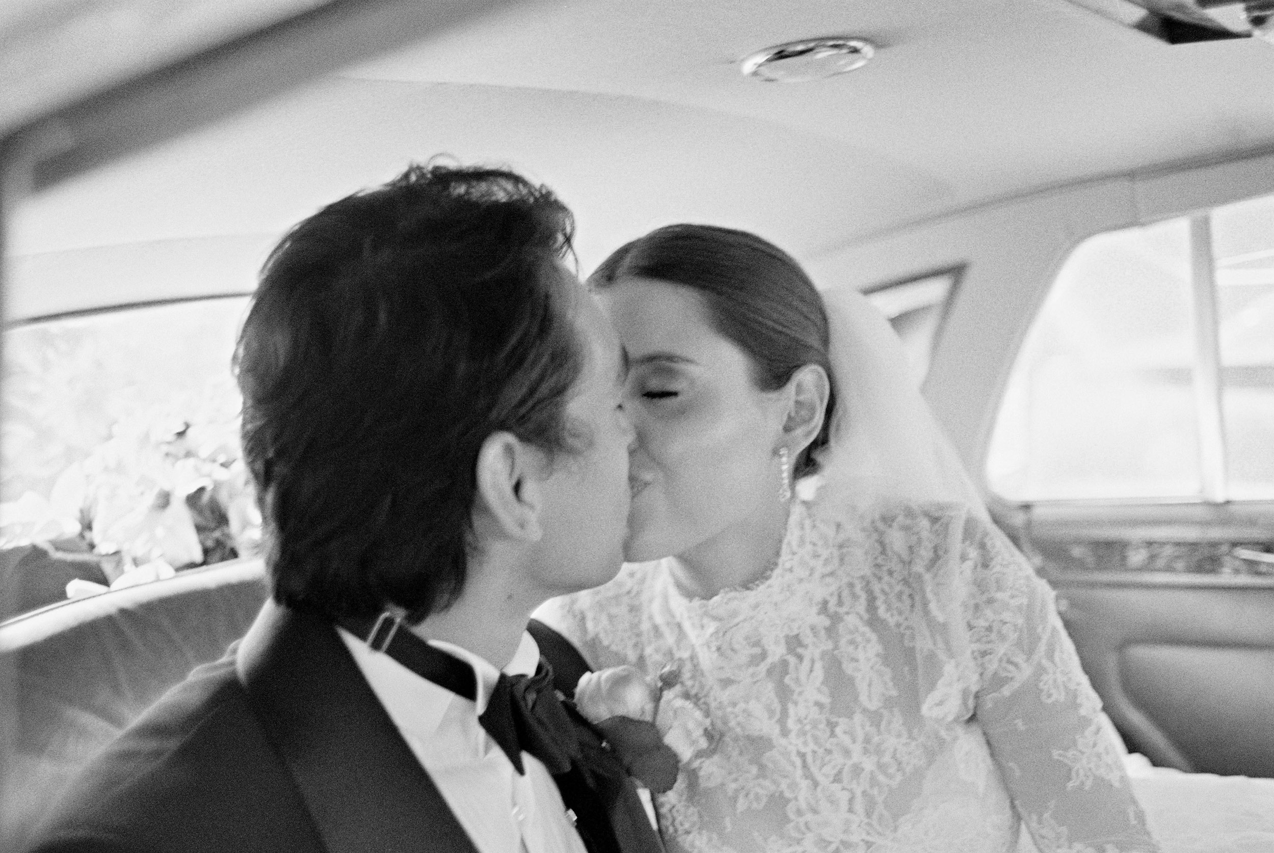 black and white 35mm film photograph, view through the open car door. A couple lean in together with the bouquet, city street inside a Rolls-Royce car.